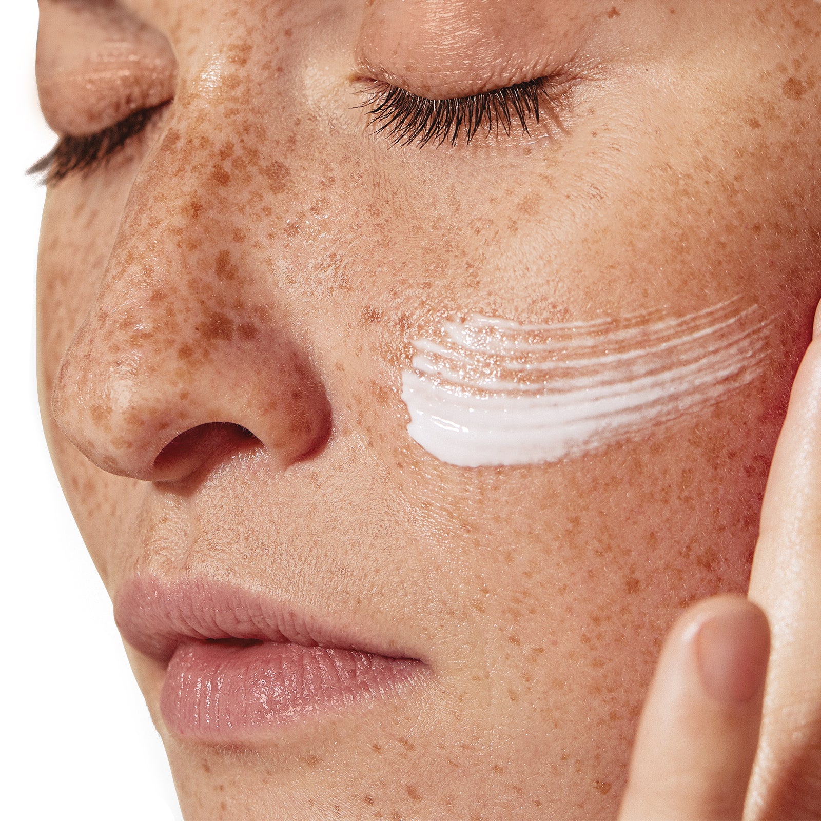 Close-up of a woman applying cream to her face with visible freckles.